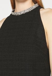 Gros plan d'une robe noire texturée sans manches avec un col montant orné d'une bande de strass clairs scintillants.
