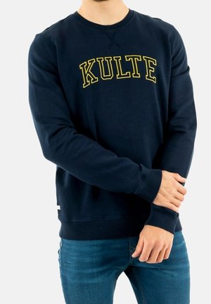 Mężczyzna w granatowej bluzie z żółtym napisem "KULTE", w niebieskich dżinsach, stojący z rękami skrzyżowanymi na talii.