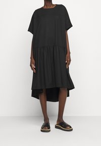 Woman wearing a loose black short-sleeve dress with a high-low hem and black platform sandals, standing against a plain white background.