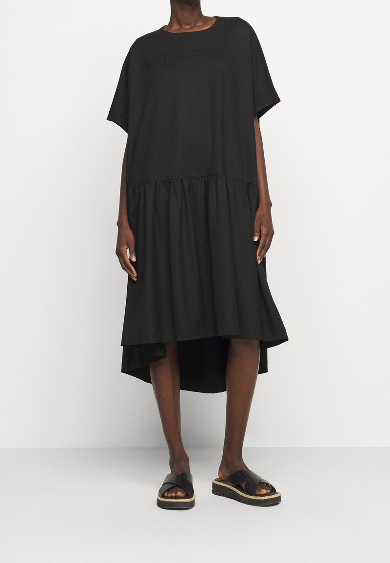 Woman wearing a loose black short-sleeve dress with a high-low hem and black platform sandals, standing against a plain white background.