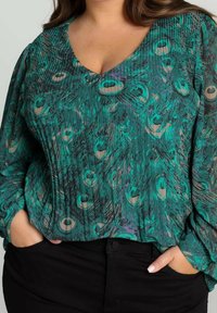 Femme portant un chemisier vert à motif de plumes de paon avec des manches longues en tulle et un pantalon noir, mains dans les poches.