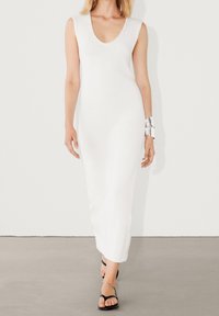 Robe blanche en maille sans manches avec un col en V profond et une longueur jusqu'au sol. Texture lisse avec une coupe décontractée. Sandales noires minimalistes à fines lanières.