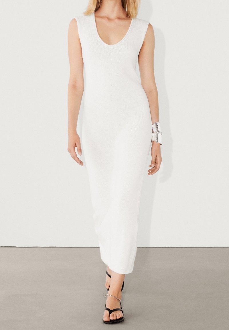 Robe blanche en maille sans manches avec un col en V profond et une longueur jusqu'au sol. Texture lisse avec une coupe décontractée. Sandales noires minimalistes à fines lanières.