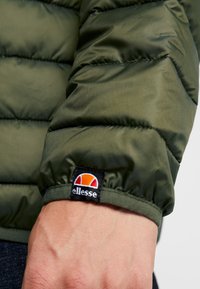 Mâneca unei jachete puffer verzi cu textură matlasată. Prezintă un etichetă neagră cu un logo alb și portocaliu la manșetă. Material neted și strălucitor.