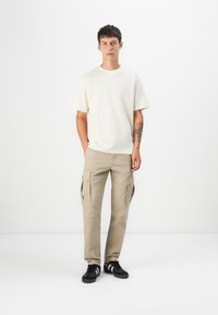 Beige katonai nadrág oldalsó zsebekkel, egy krém színű, rövid ujjú kötött pólóval párosítva. Fekete sportcipő fehér csíkkal.