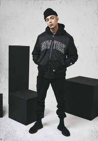 Chaqueta bomber negra con "NEW YORK" impreso en blanco, superpuesta a una sudadera con capucha negra. Pantalones cargo negros y zapatillas negras completan el atuendo.