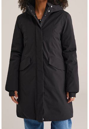 Manteau d'hiver long noir avec capuche, poches avant à rabat et boutons pression, porté par-dessus un jean bleu.