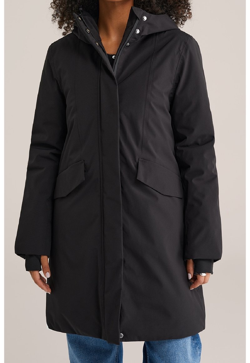 Manteau d'hiver long noir avec capuche, poches avant à rabat et boutons pression, porté par-dessus un jean bleu.