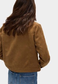 Giacca in suede marrone con una vestibilità aderente, dotata di orlo arrotondato, dettagli di cucitura minimali, indossata con jeans a vita alta.