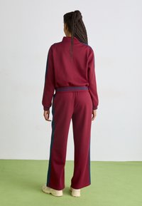 Bordeaux tracksuit med blå sidestropper, med høj krave og elastisk talje. Blød stoftekstur med en afslappet pasform.