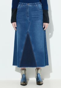 Falda maxi de mezclilla que presenta una combinación de paneles en tonos de azul claro y oscuro, con una abertura frontal y un dobladillo deshilachado, combinada con botas metálicas hasta el tobillo.