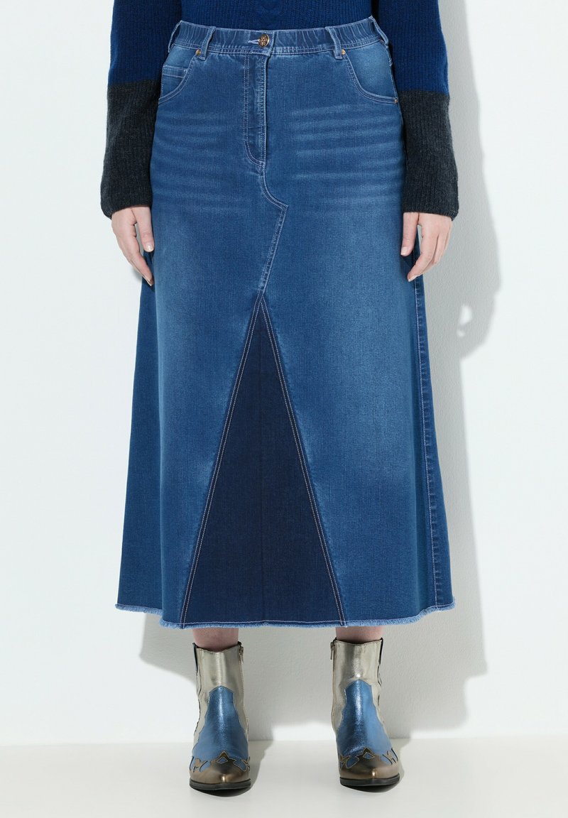Falda maxi de mezclilla que presenta una combinación de paneles en tonos de azul claro y oscuro, con una abertura frontal y un dobladillo deshilachado, combinada con botas metálicas hasta el tobillo.