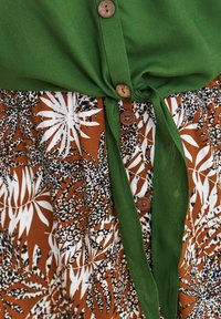 Zielona bluzka z detalami w postaci guzików, związana w talii, nałożona na brąskową spódnicę z białymi i czarnymi liśćmi. Materiał teksturowany.