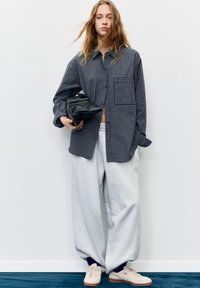 Checkered dark blue shirt, gray sweatpants, and beige sneakers. The shirt has a pocket and button closure; a black bag is held in one hand.