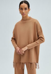 Camel-kleurige gebreide trui met geribbelde boorden en hoge halslijn, met een losse pasvorm en franjes aan de zoom. Gladde textuur.