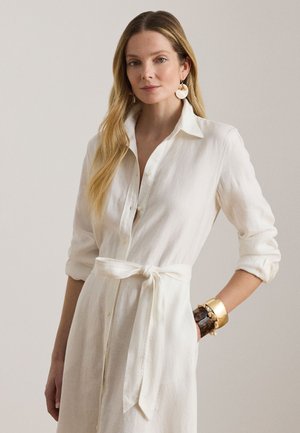 Ženska z dolgimi svetlo rožnatimi lasmi, oblečena v belo obleko z gumbi in pasom, velike okrogle uhane ter več zlatih in temnih zapestnic na zapestju.