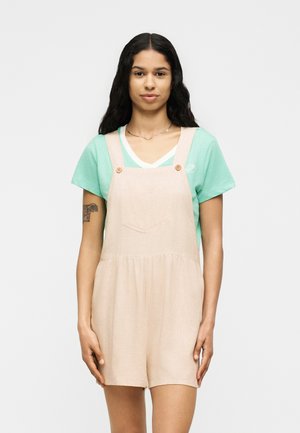 Jeune femme aux longs cheveux foncés portant un t-shirt vert menthe sous une salopette courte en lin beige, debout devant un fond blanc uni.