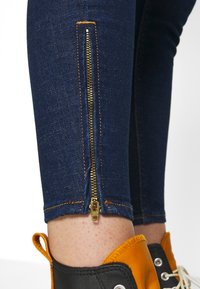 Jeans de mezclilla oscura con un cremallera dorada visible en el tobillo, con costuras en naranja y un ajuste ceñido. Combinados con zapatillas negras y naranjas.
