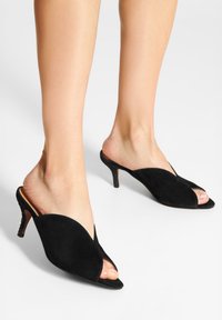 Svart mocka öppen tå mules med en v-formad utskärning och en avsmalnande klack. Har slät textur och en minimalistisk stil.