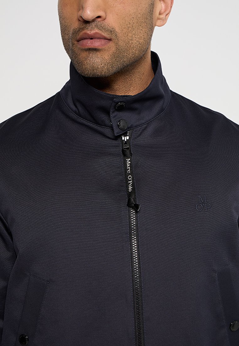 Mann in einer dunkelblauen Jacke mit Reißverschluss und hohem Kragen. Der Reißverschluss hat einen „Marc O'Polo“-Zip-Puller und das gestickte „M OP“-Logo auf der Brust.