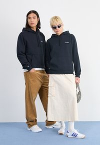 Kaks mudelit, kellel on seljas mustad Salomoni kapuutsid; üks pruunides sametpükstes, teine kreemjas cargo-seelikus ja päikeseprillides, hoides mustrilist kotti.