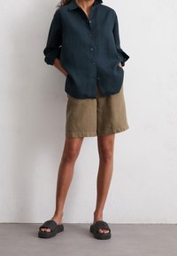 Camicia di lino verde scuro con bottoni e tasca, abbinata a pantaloni corti di lino color khaki e sandali neri slip-on con texture.