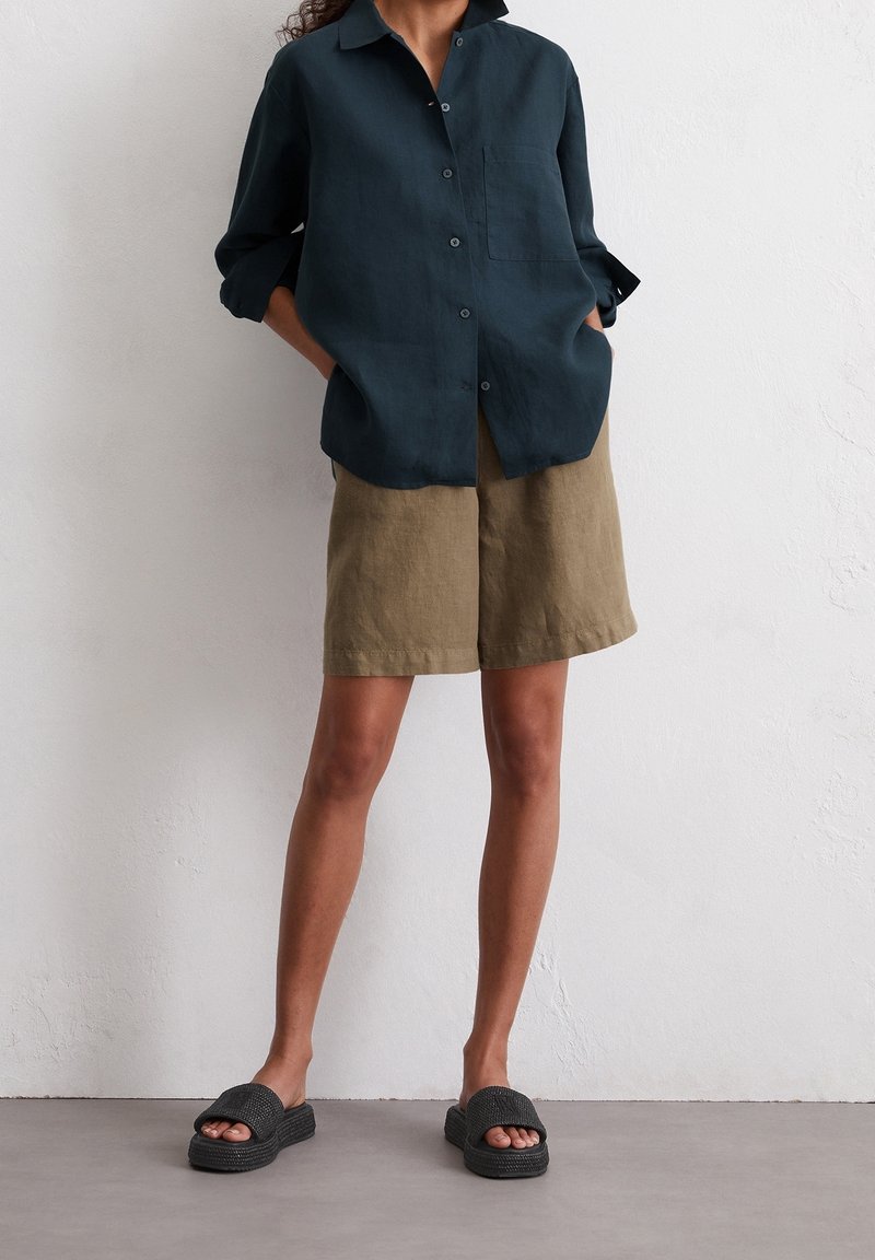 Camicia di lino verde scuro con bottoni e tasca, abbinata a pantaloni corti di lino color khaki e sandali neri slip-on con texture.