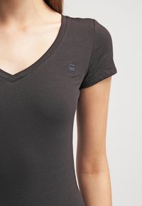 Mörkgrå tajt t-shirt med v-ringning, korta ärmar och en liten logotyp tryckt på bröstet. Tillverkad av mjukt, elastiskt material.