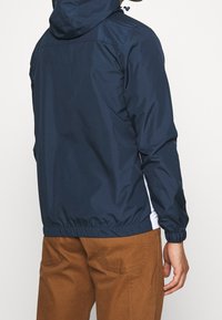 Veste légère bleu marine avec capuche, manches et ourlet élastiques ; détails latéraux en blanc ; texture de tissu lisse. Fini avec une légère brillance.