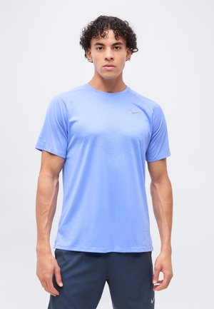 Jonge man met krullend haar die een lichtblauw Nike sportshirt met korte mouwen en donkere korte broek draagt, staand tegen een effen achtergrond.