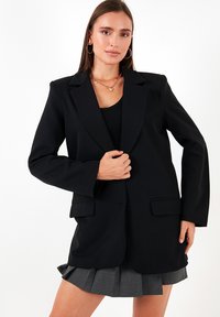Sacou negru cu o croială ajustată, rever cu decupaj, două buzunare frontale și mâneci lungi. Purtați peste un top negru și o fustă plisată gri.