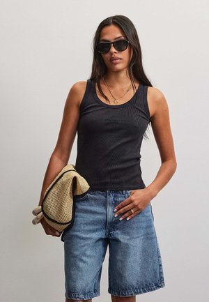Kvinde iført sort tanktop, denim bermudashorts og solbriller, holder en stråhat og handsker, står mod en ensfarvet baggrund.