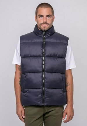 Homme aux cheveux courts portant un gilet matelassé bleu marine sur une chemise blanche à manches courtes et un pantalon vert olive, debout devant un fond gris.