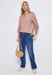Hellrosa Bluse mit Notch-Kragen und hochgekrempelten Ärmeln, kombiniert mit blauen geraden Jeans. Die Accessoires bestehen aus einer beige Umhängetasche und weißen Sneakern.