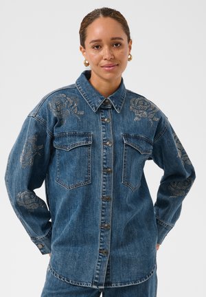 Femme portant une chemise en jean bleu avec des motifs de roses métalliques sur les épaules et les manches, boucles d’oreilles dorées, debout devant un fond clair et uni.