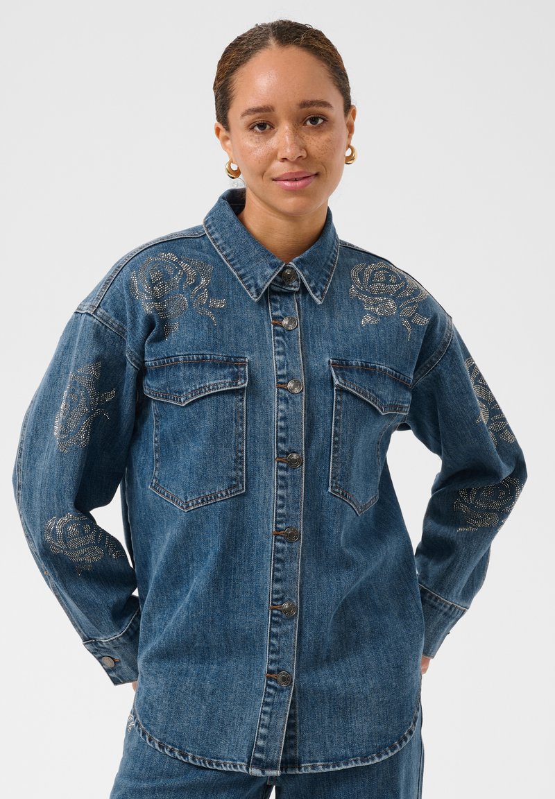 Femme portant une chemise en jean bleu avec des motifs de roses métalliques sur les épaules et les manches, boucles d’oreilles dorées, debout devant un fond clair et uni.