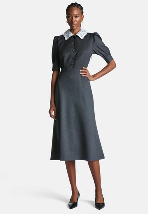 Femme portant une robe midi gris foncé avec des manches bouffantes, des boutons dorés, un col en dentelle blanche, des talons noirs et des boucles d'oreilles créoles dorées, debout avec assurance.