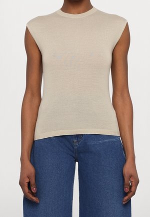 Person trägt ein ärmelloses beiges Stricktop kombiniert mit hoch taillierten blauen Jeans vor einfarbigem Hintergrund.