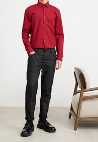 Rotes Hemd mit Knöpfen und langen Ärmeln, schwarze, eng anliegende Hose und glänzende schwarze Schuhe mit dicken Sohlen. Schlichtes, strukturiertes Design.