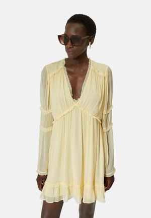 Femme portant une robe jaune clair à manches longues avec des détails à volants et des lunettes de soleil grandes et foncées, debout devant un fond uni.