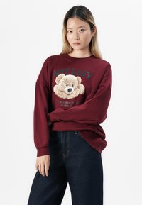Young woman with long hair wears maroon sweatshirt featuring a teddy bear and text, paired with dark blue jeans, standing against white background.