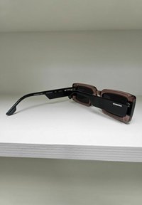 Lunettes de soleil rectangulaires avec des verres teintés. La monture présente un avant translucide marron foncé et des branches noires mat, étiquetées "KOMONO."