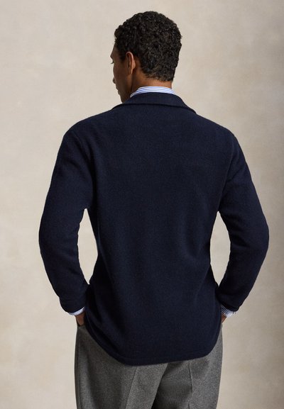 Uomo con capelli corti e ricci che indossa un maglione nero sopra una camicia a righe blu e bianca con colletto e pantaloni grigi, mani in tasca, di spalle.