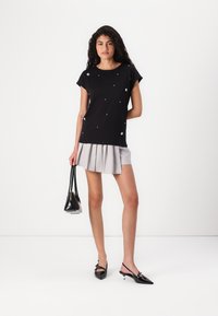 Black short-sleeve top with floral embellishments, paired with a light gray pleated skirt. Model holds a black handbag and wears pointed shoes.