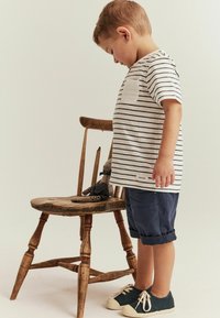 Randig t-shirt med bröstficka, blå rullade shorts och mörka sneakers. En trästol håller en mjukisdjur. Neutral bakgrund.