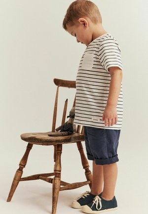 Stribet t-shirt med brystlomme, blå rullede shorts og mørke sneakers. En træstol holder en blød legetøjsfigur. Neutral baggrund.
