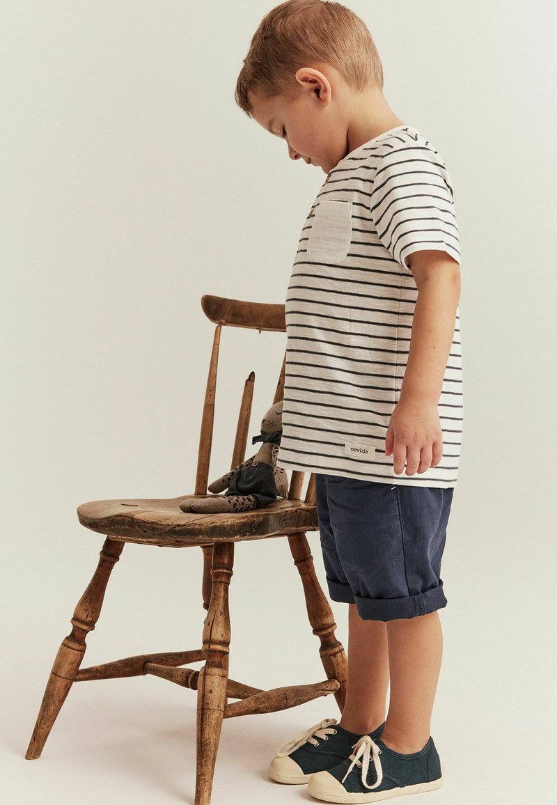 Randig t-shirt med bröstficka, blå rullade shorts och mörka sneakers. En trästol håller en mjukisdjur. Neutral bakgrund.