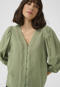 Blusa verde con maniche a sbuffo corte, scollo a V e chiusura con bottoni. Presenta un bordo in pizzo e un tessuto testurizzato per un tocco di dettaglio in più.