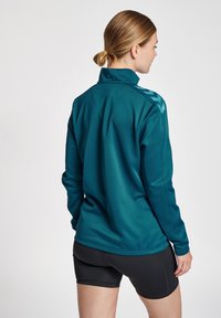 Giacca sportiva teal con colletto alto, maniche lunghe e accenti a forma di chevron sulle spalle, abbinata a pantaloni corti neri. Texture liscia.
