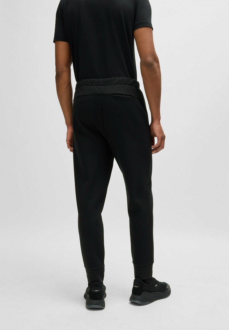 Pantalones joggers negros con cintura elástica, ajuste ajustado y puños de canalé en los tobillos, combinados con zapatillas negras. Diseño simple y funcional.